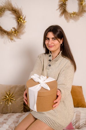Young beautiful European woman with dark hair holding a Christmas gift box with a bow, in a bright room with a Christmas tree, with a bed, garlands, her face is blurred, the box is in focusの写真素材