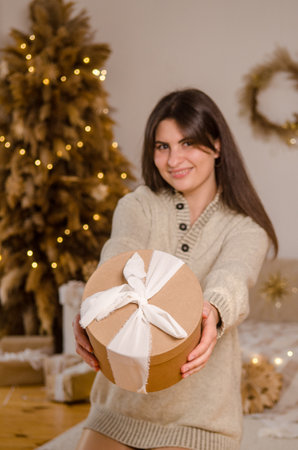 Young beautiful European woman with dark hair holding a Christmas gift box with a bow, in a bright room with a Christmas tree, with a bed, garlands, her face is blurred, the box is in focusの写真素材
