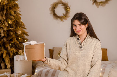 Young beautiful European woman with dark hair holding a Christmas gift box with a bow, in a bright room with a Christmas tree, with a bed, garlandsの写真素材