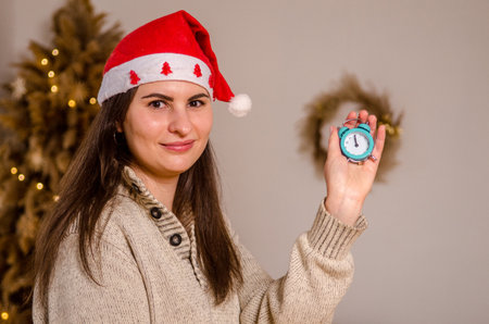 Young beautiful woman in a sweater with dark hair in red santa hat with stars, against background of bed and Christmas tree, in bright room, holds retro alarm clock with time of midnight in her handの写真素材