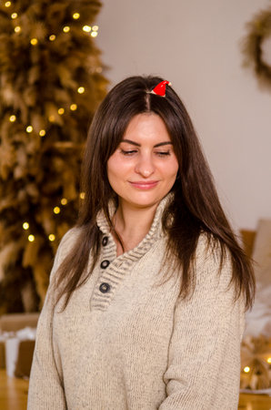 Young beautiful cheerful happy woman in a sweater and with dark hair wears a small santa hat as headdress against background of bed, garland, Christmas tree in a bright roomの写真素材
