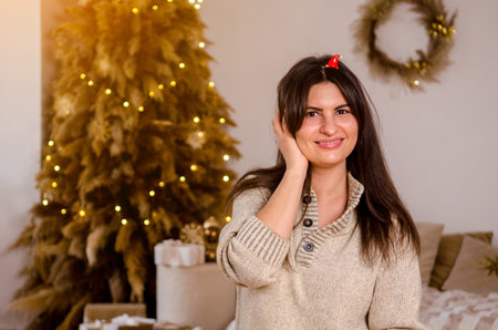 Young beautiful cheerful happy woman in a sweater and with dark hair wears a small santa hat as headdress against background of bed, garland, Christmas tree in a bright roomの写真素材