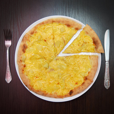 Delicious fresh round cheese pizza with pear, pineapples on black table, surface, on white plate, fork and knife nearby, top viewの写真素材