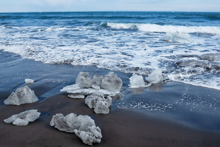 Beautiful winter sunny landscape of the Pacific Ocean coast. Black volcanic sand and ice blocks that...
