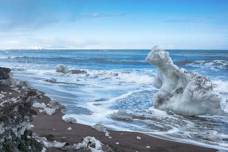 Beautiful winter sunny landscape of the Pacific Ocean coast. Black volcanic sand and ice blocks that...