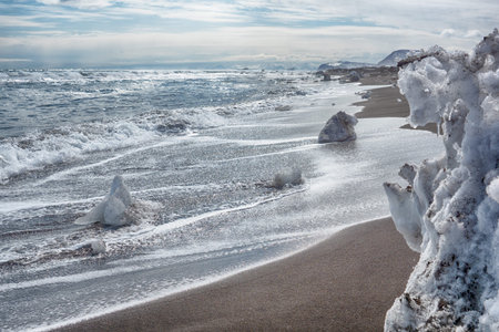Beautiful winter sunny landscape of the Pacific Ocean coast. Black volcanic sand and ice blocks that...