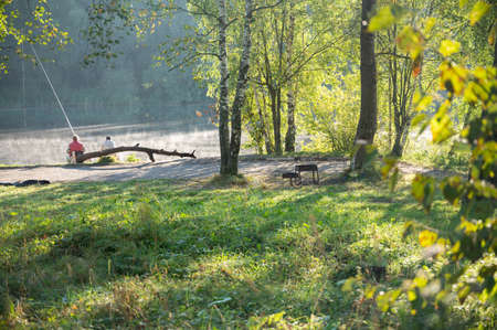 two fishermen are sitting on the shore near the lake with long rods fishing, talking, enjoying.Around the beautiful nature, forest, glades.There is a barbecue in the distance.Foggy morning on the pondの写真素材