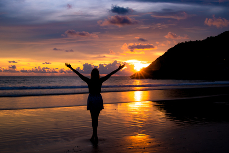 Silhouette of woman with hands up while standing on the sea beach at sunset.の写真素材