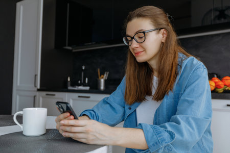 woman sitting in kitchen with smartphone, texting, browsing online services, working or studying remotely from homeの写真素材