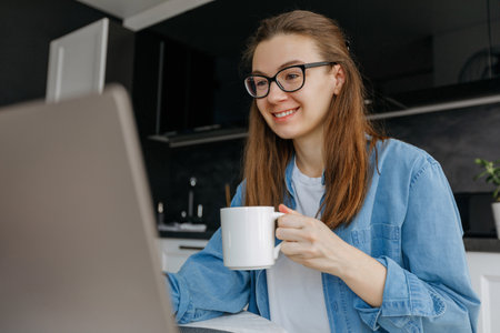 Relaxed female freelancer relaxing sitting with cup of coffee in front of laptop, working or studying remotely from home, drinking from mug taking a breakの写真素材