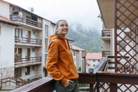 Portrait of a beautiful young woman in an orange sweatshirt standing on a balcony overlooking the mountainsの写真素材