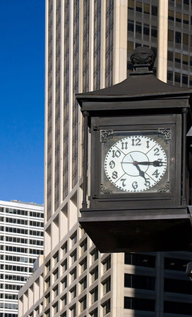 Old-fashioned iron street clock mounted on the wall in front of modern  skyscrapers in New York downtown financial district (Wall Street) on the blue sky background.  The time is fifteen after five.の写真素材