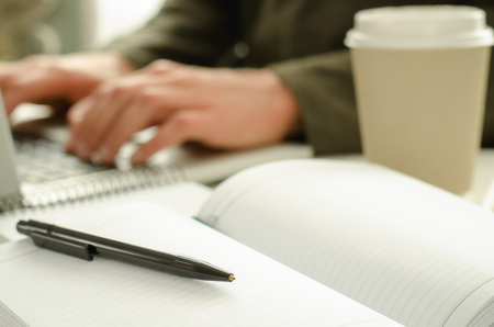 Open notepad with black pen on work-table. Paper cup of coffee, office stuff, laptop and working man on backgroundの写真素材