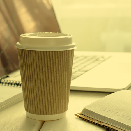 Paper cup of coffee on work-table with office stuff, notepad, laptop, pen and coffee cupの写真素材