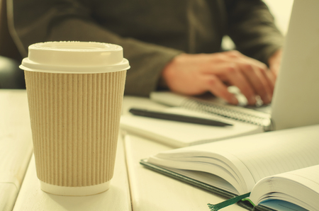 Paper cup of coffee on work-table with office stuff, notepad, laptop, pen, coffee cup and working man on backgroundの写真素材
