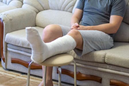 The guy is sitting on the couch in a cast. The leg is in a cast. Broken leg.の写真素材