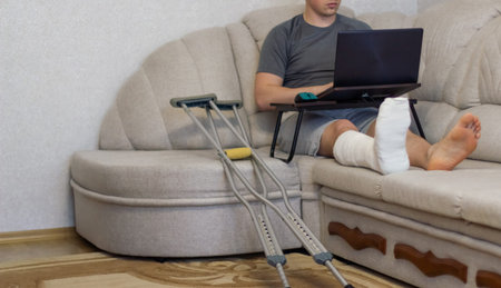 Man is lying on sofa and works at the computer with bandage on leg.