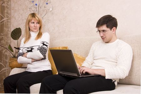 Woman is angry with man for working at laptop. Female takes offence at male. Man is sitting on the sofa and he is busy.の写真素材