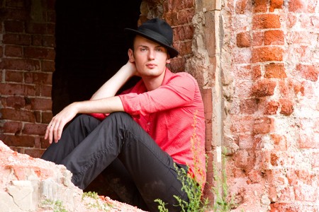 Elegant fashion man model wear red shirt and hat near bare walls. Handsome young man look. Young casual man portrait wear a hatの写真素材