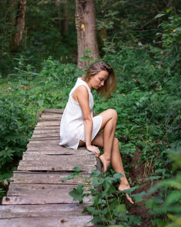 Woman in white short dress in summer forest sits barefoot on wooden bridgeの写真素材