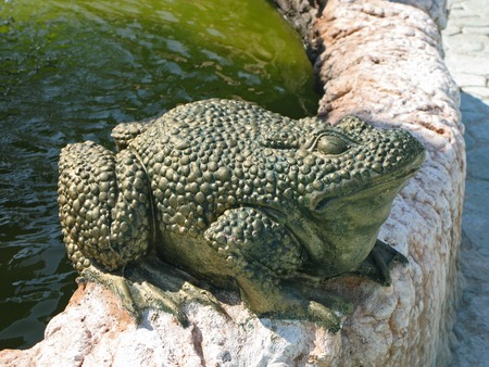 The sculpture is a large green frog on the stone curb of the fountainの写真素材