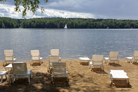 Empty plastic loungers on a sandy beach on the shore of a lake with sailboatの写真素材
