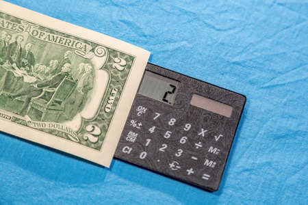 Part of a two dollar American bill and a compact digital calculator on a fabric blue backgroundの写真素材