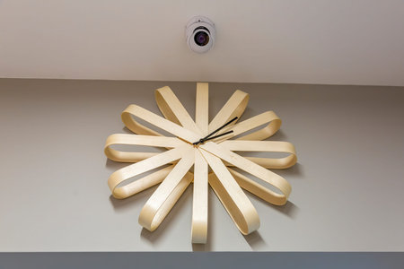 Mechanical wooden clock with black arrows and surveillance camera on beige interior wall