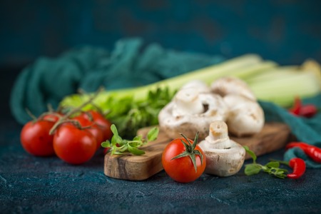 Fresh mushrooms champignons on a wooden cutting board, parsley greens, celery stalks, cherry tomatoes and hot red chili pepper on a dark blue backgroundの写真素材
