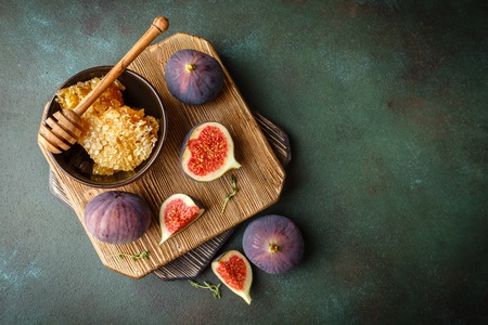 Juicy fresh whole fig fruits and one cut figs and bowl of honey in honeycombs on wooden cutting board on dark green background. Top viewの写真素材