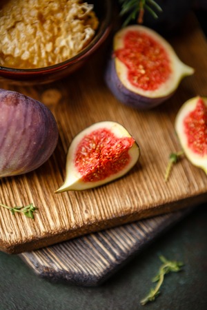Juicy fresh whole fig fruits and one cut figs and bowl of honey in honeycombs on wooden cutting board on dark green backgroundの写真素材