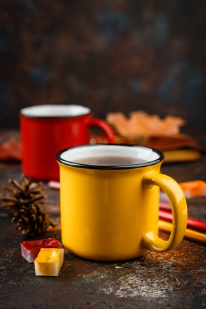 Yellow and red colored enamel cups of tea, watercolors in cuvettes, colored pencils, autumn maple leaves and bumps on a brown backgroundの写真素材