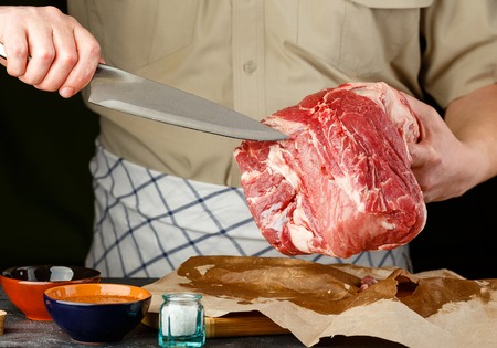 Man cuts large piece of pork meat on wooden cutting board with knife. Close up.の写真素材