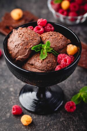 Three brown chocolate ice cream balls with raspberry and mint leaves in black bowl on dark backgroundの写真素材