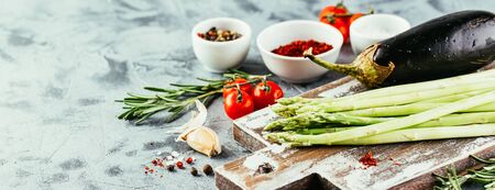 Fresh asparagus, tomatoes, eggplant, garlic, pepper and salt with sprigs of rosemary on wooden cutting board on gray background. Long wide bannerの写真素材