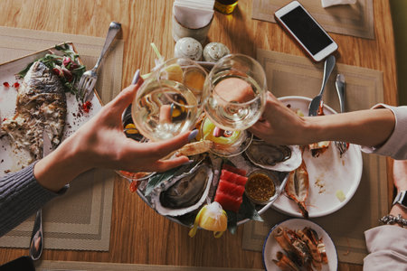 Two young woman clinking glasses with wine over served table with food in cafe or restaurant. Close-up hands with wineglassesの写真素材