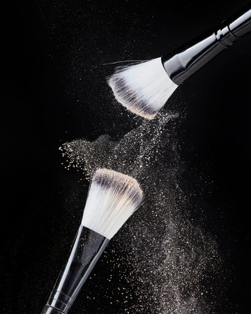 Two professional black makeup brushes with beige powder in motion and loose powder particles...
