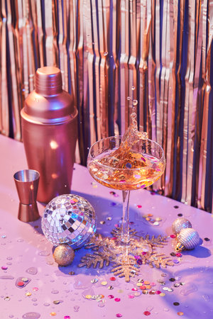 Splash in champagne glass, cocktail glass, shaker, and jigger on pink table with Christmas decorationsの写真素材