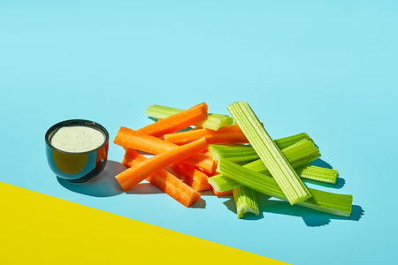 Raw carrot pieces and celery stalks with sauce on color background