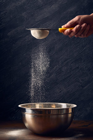 Hand sifting wheat flour through sieve at table on black background, close-up