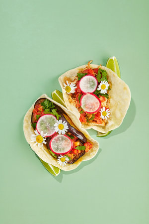 Tacos on corn tortillas with chicken and vegetable filling decorated with daisy flowers on green background with lime wedgesの写真素材