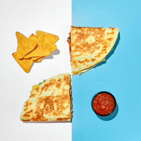 Two halves of quesadilla with chicken, cheese, vegetables, herbs and salsa sauce and nachos corn chips on colored backgroundの写真素材