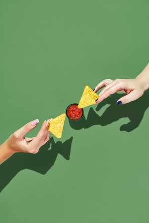 Two female hands reaching towards each other with nacho chips over salsa sauce bowlの写真素材