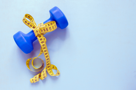 Fitness and weight loss concept. Blue dumbbell with bow of yellow centimeter on a blue background. Top view.の写真素材