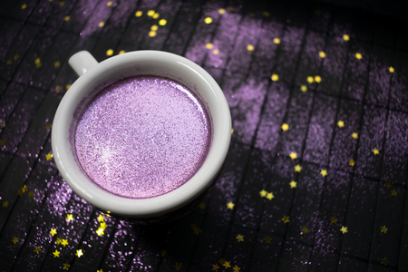 Cup of coffee with purple glitter on dark background with golden stars. Glitter cappuccino, sparkly shine  gold espresso, coffee with sparkle. Trendy drink, original concept. Holiday party background.の写真素材