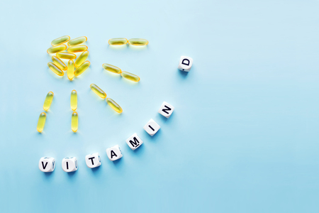 Yellow capsules in the form of the sun with rays and the word vitamin D from white cubes with letters on a blue background. VITAMIN D word for healthy and medical concept. Sunshine vitamin health benefitsの写真素材