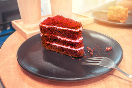 A piece of red velvet cake on a brown round plate with forks and crumbs. A serving of red velvet...