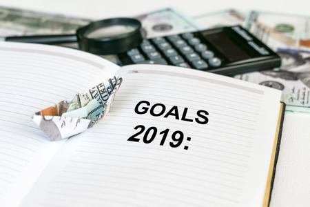 2019 goals text in open notebook with dollar banknote origami ship and calculator, money and magnifier in the background. Business financial  Conceptの写真素材