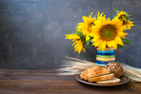 Autumn harvest, natural products, Greeting card concept. Still life with beautiful sunflowers in vase, sliced white bread on clay plate, wheat spikelets on wooden rustic background.の写真素材
