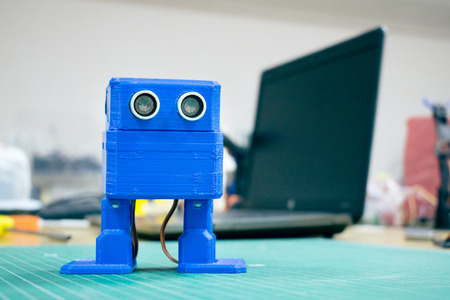 3D printed Funny dancing blue robot on the background of devices and laptop. Robot model printed on automatic three dimensional 3d printer. Additive manufacturing and robotic automation technology.の写真素材